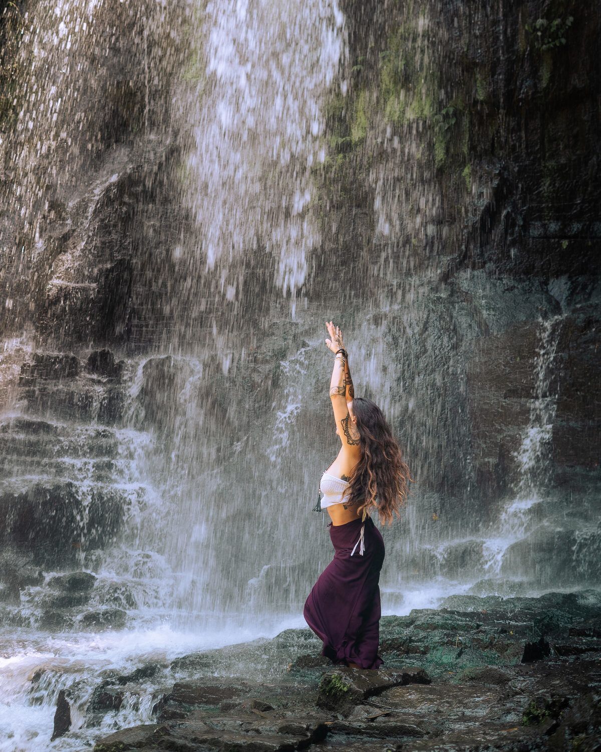 TIHANE beneath a waterfall, arms raised